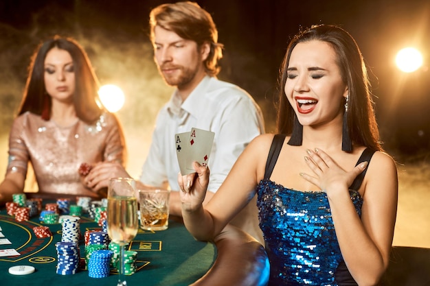 도박장에서 포커를 하는 우아한 사람들의 그룹입니다. 반짝이는 파란색 드레스를 입은 감성적인 갈색 머리에 집중하세요. 열정, 카드, 칩, 알코올, 주사위, 도박, 카지노 - 엔터테인먼트입니다. 위험