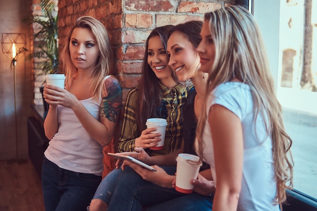 Group of female friends in casual clothes discussing while looking something on a digital tablet in a room with a loft interior.