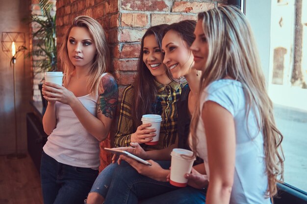 Group of female friends in casual clothes discussing while looking something on a digital tablet in a room with a loft interior.