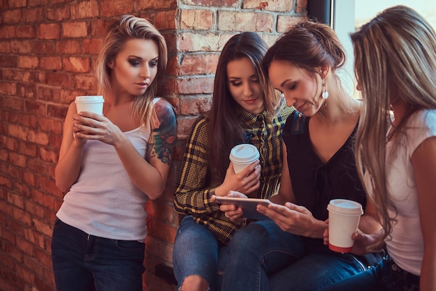 Group of female friends in casual clothes discussing while looking something on a digital tablet in a room with a loft interior.