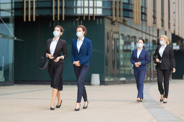 都市の建物を過ぎて一緒に歩いて、話して、プロジェクトについて議論するオフィススーツとマスクの女性マネージャーのグループ。 covidの流行の概念における完全な長さのビジネス