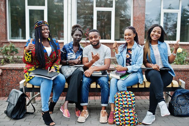 Group of five african college students spending time together on campus at university yard Black afro friends studying at bench with school items laptops notebooks