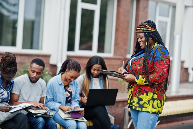 Group of five african college students spending time together on campus at university yard Black afro friends studying at bench with school items laptops notebooks