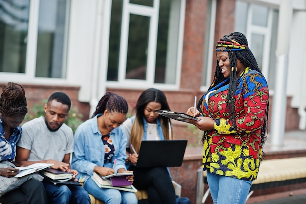 Group of five african college students spending time together on campus at university yard Black afro friends studying at bench with school items laptops notebooks