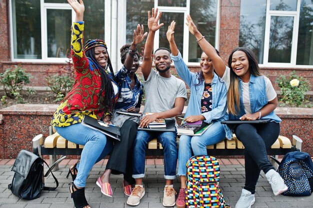 Группа из пяти африканских студентов колледжа, проводящих время вместе в кампусе на университетском дворе Черные афро друзья учатся на скамейке со школьными принадлежностями, ноутбуками, ноутбуками