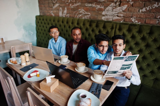 Группа из четырех мужчин из Южной Азии позирует на деловой встрече в кафе. Индийцы вместе работают с ноутбуками, используя различные гаджеты, разговаривают и смотрят в меню