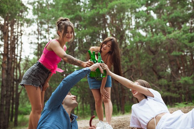 Group of friends clinking beer bottles during picnic in summer forest
