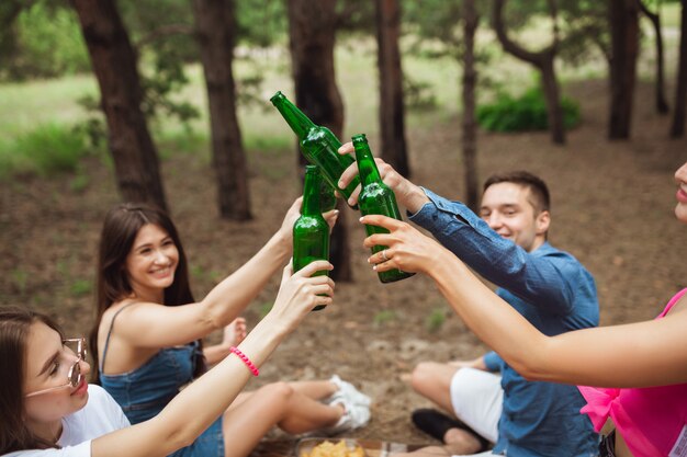 Group of friends clinking beer bottles during picnic in summer forest