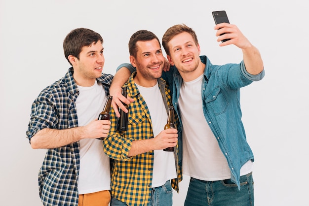 Group of friends holding beer bottle taking selfie on mobile phone against white backdrop
