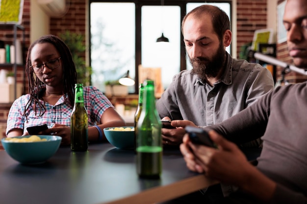 Group of friends sitting at table in living room while browsing social media on smartphones. Diverse people with modern wireless touchscreen devices sitting at home while enjoying free time.