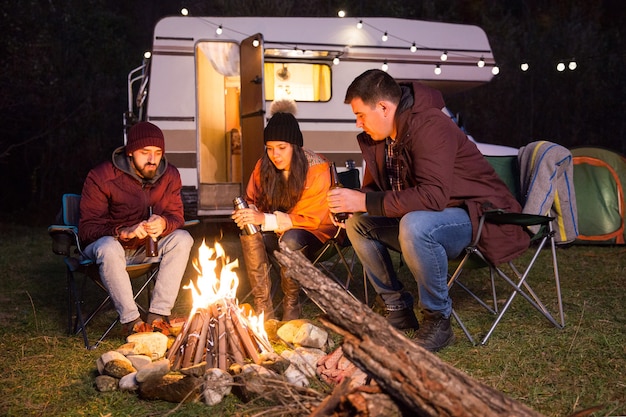 Group of friends sitting together around camp fire in a cold night of autumn in the mountains. Retro camper van with light bulbs.