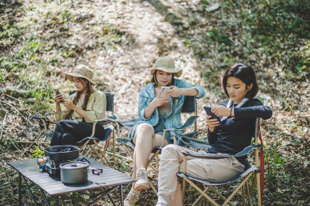 Group of girl friends sitting on camping chair and use their smartphone ignoring each other while camping in park