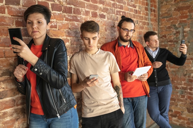 Group of happy caucasian young people standing behind the brick wall. sharing a news, photos or videos from smartphones or tablets, playing games and having fun. social media, modern technologies.