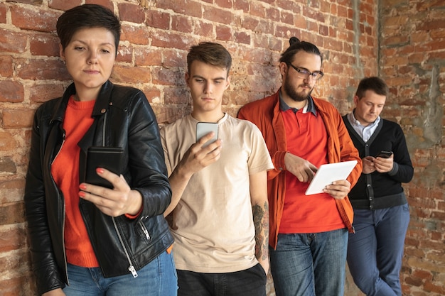 Group of happy caucasian young people standing behind the brick wall. Sharing a news, photos or videos from smartphones or tablets, playing games and having fun. Social media, modern technologies.