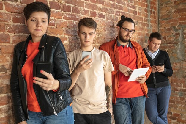 レンガの壁の後ろに立っている幸せな白人の若者のグループ。スマートフォンやタブレットからのニュース、写真、ビデオの共有、ゲームのプレイ、楽しみ。ソーシャルメディア、最新テクノロジー。