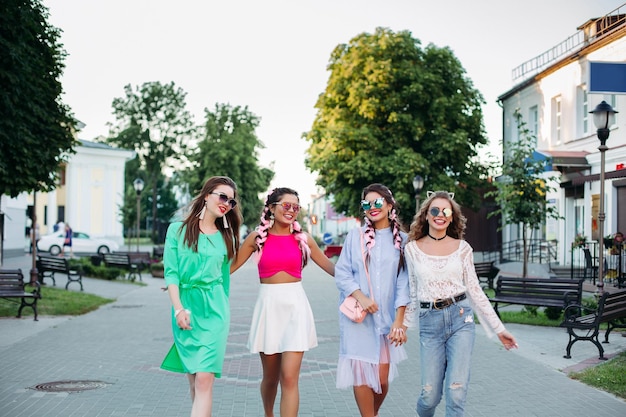 Group of happy fashionable women in sunglasses walking at street embracing smiling Pretty stylish best girlfriends in colorful clothes with pink brides after shopping Street style look