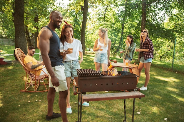 Group of happy friends having beer and barbecue party at sunny day. Resting together outdoor in a forest glade or backyard. Celebrating and relaxing, laughting. Summer lifestyle, friendship concept.