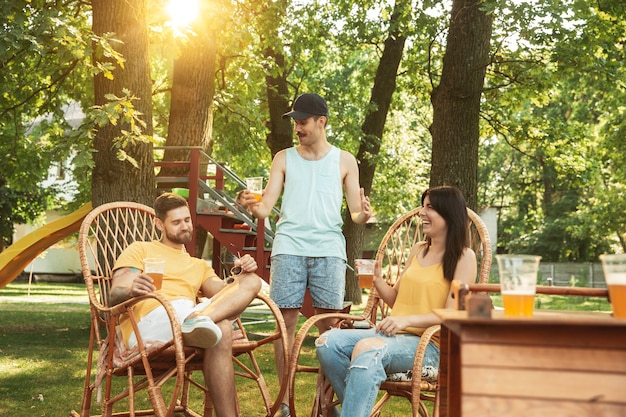 Group of happy friends having beer and barbecue party at sunny day. Resting together outdoor in a forest glade or backyard