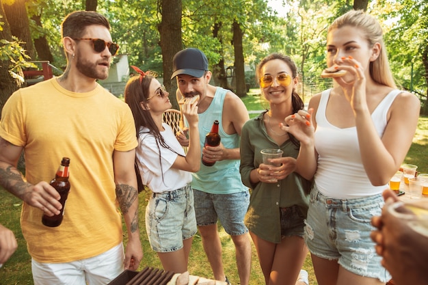 Group of happy friends having beer and barbecue party at sunny day. Resting together outdoor in a forest glade or backyard