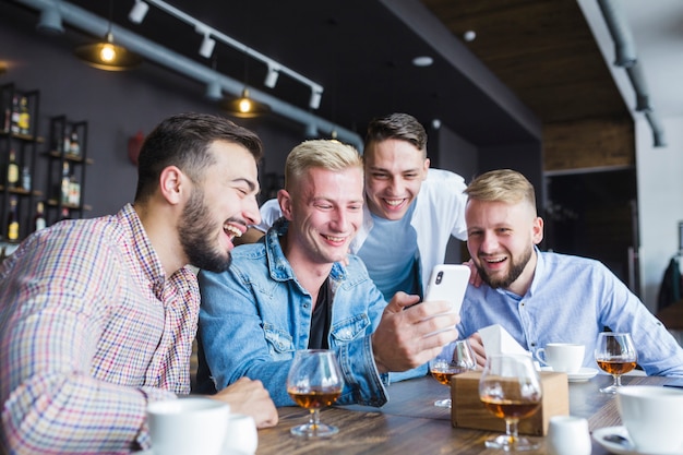 Group of happy friends looking at smartphone sitting at restaurant