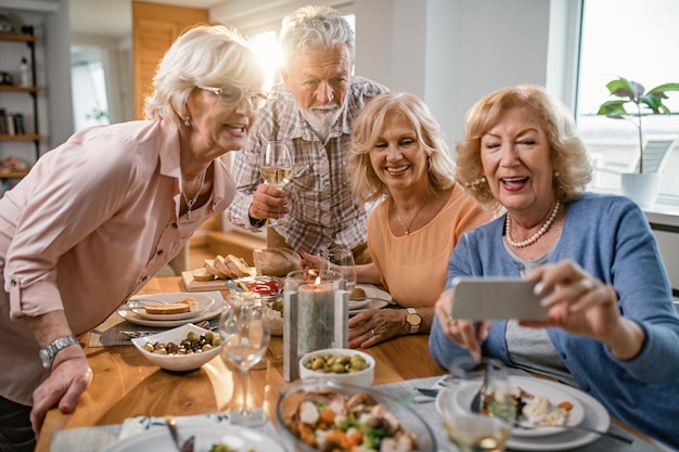 Группа счастливых зрелых людей, делающих селфи с мобильным телефоном, обедая вином и едя за обеденным столом