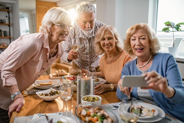 Group of happy mature people taking selfie with mobile phone while dining wine and eating at dining table