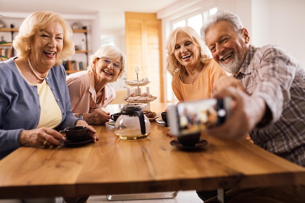 Group of happy senior friends taking selfie while gathering on afternoon tea at home