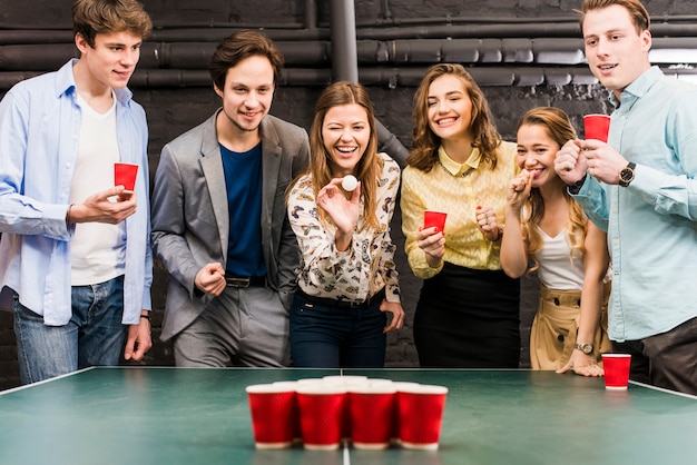 Gruppo di amici sorridenti felici che godono del gioco del pong della birra sulla tavola nella barra