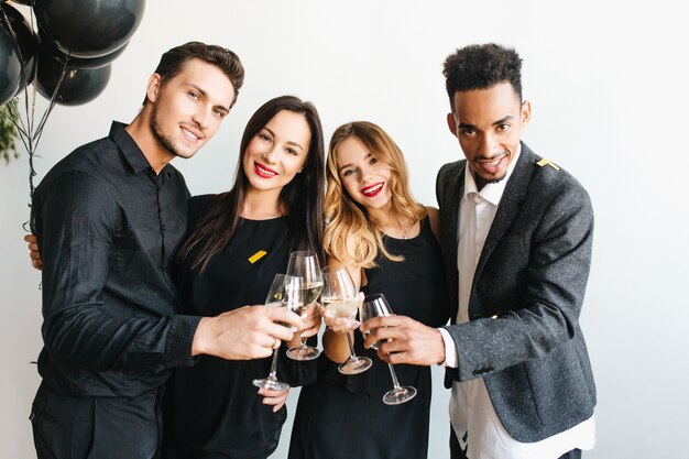 Group of joyful young people in trendy attires raising glasses with champagne