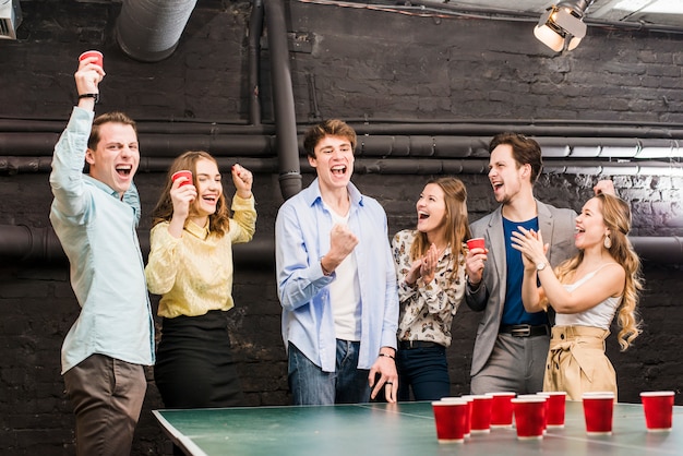 Group of laughing friends enjoying beer pong on table