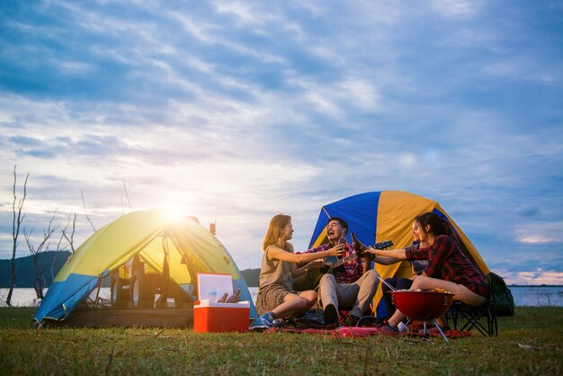 남자와 여자의 그룹 배경에서 텐트와 호수에서 캠핑 피크닉과 바베큐를 즐길 수 있습니다. 젊은 혼합 인종 아시아 여자와 남자. 맥주 병을 토스트하고 응원하는 젊은 사람들의 손에.