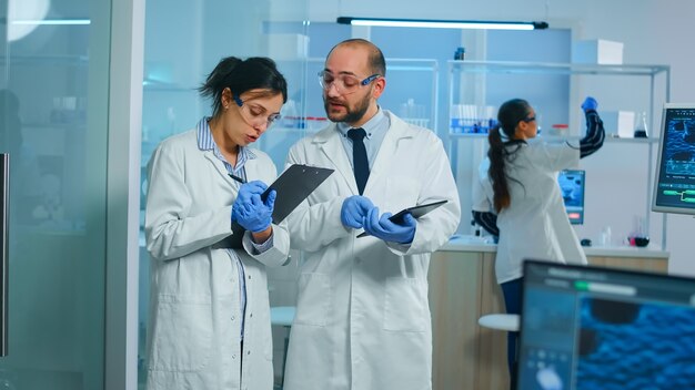 Group of medical researchers discussing about vaccine development standing in equipped lab pointing on tablet and taking notes