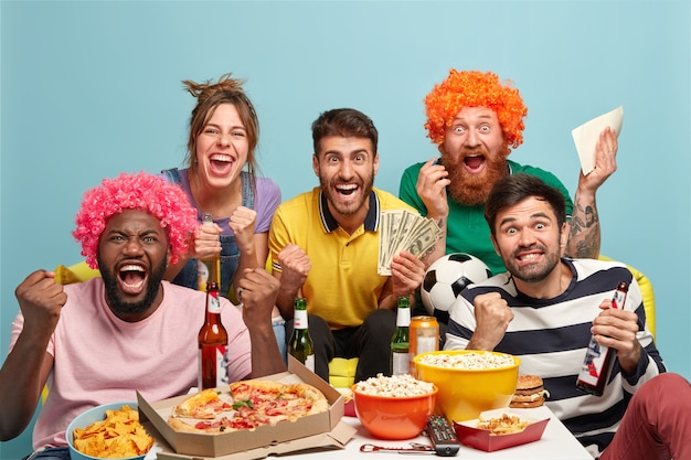 Group of mixed race best friends watch football match with excitement, shout for favorite team, make sport betting on money, clench fists, eat pizza, popcorn, drink beer, celebrate goal, cheer up