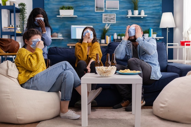 Group of multiracial friends watching horror show on tv enjoying spending time together wearing face mask to prevent infection with covid 19, during global pandemic having fun sitting on couch