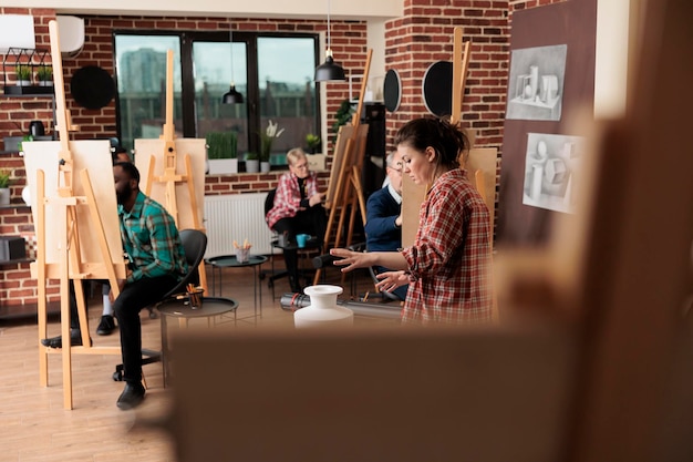 無料写真 グループアートクラスで絵を描くことを勉強するイーゼルに座っているさまざまな年齢の人々のグループ。静物スケッチのテクニックを生徒に説明する若い女性教師。大人向けの社会活動