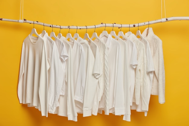 Бесплатное фото Группа белой простой одежды, висящей на вешалке или рельсе. минималистичная концепция. одежда для женщин, изолированных на желтом фоне.