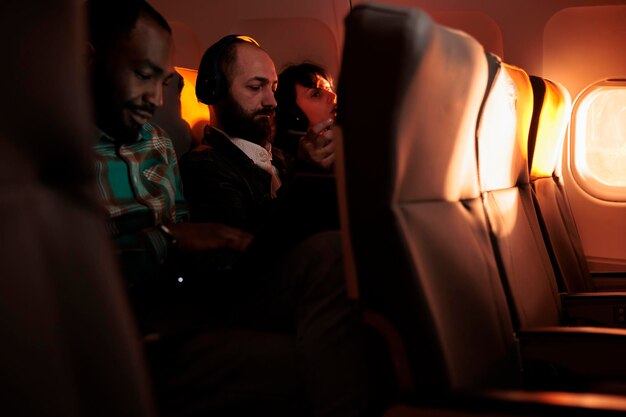 Группа пассажиров, путешествующих в пункт назначения на самолете, использующих компьютер или мобильный телефон в полете во время заката. Летим эконом-классом с людьми, чтобы отправиться в отпуск или в командировку.