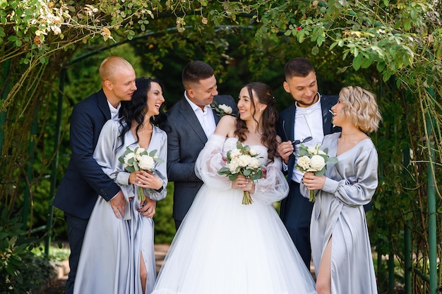 Group portrait of couples bride with groom and bridesmaids with groomsmen posing in the garden near the arch smiling looking at each other Wedding day celebration outdoors ceremony