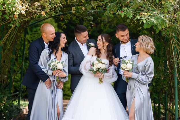 Group portrait of couples bride with groom and bridesmaids with groomsmen posing in the garden near the arch smiling looking at each other Wedding day celebration outdoors ceremony