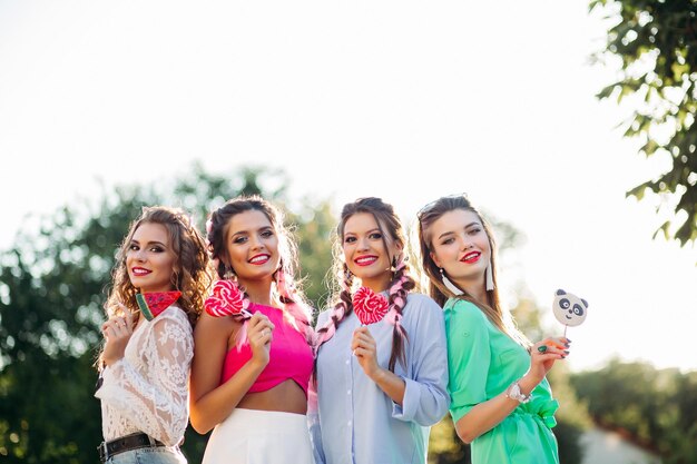 Group of pretty and fashionable girls wearing in trendy clothes with colorful braid and professional makeup, walking at street after shopping. Women holding candies heart on stick and smiling.