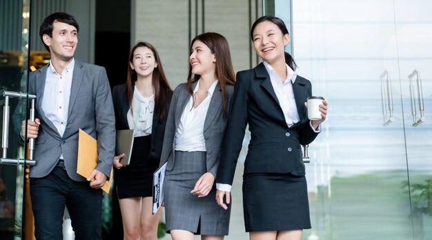 영리한 젊은 아시아 사업가와 여성 정장을 입고 걷는 그룹은 자신감과 행복으로 현대적인 사무실 입구를 통과합니다.