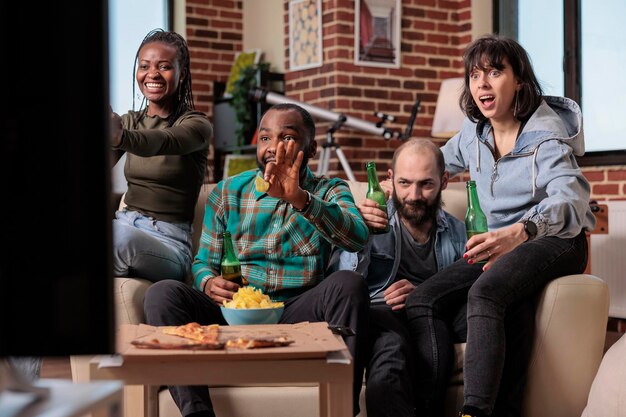 Group of supporters watching football game tournament on sport tv channel, cheering in front of television to score goal on pitch. Looking at soccer match championship and drinking beer for fun.