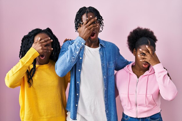 Group of three young black people standing together over pink background peeking in shock covering face and eyes with hand looking through fingers with embarrassed expression