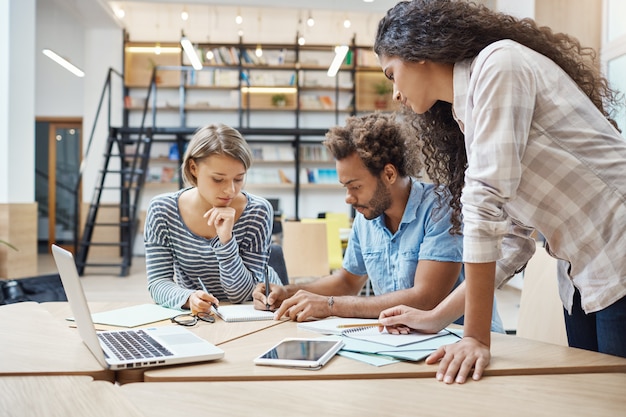 Group of three young multi-ethnic successful business people sitting in coworking space, talking about new project of competitor team, making plans to bypass their project.