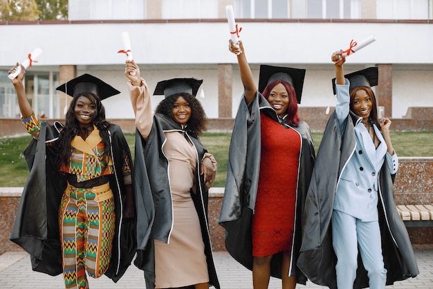 Free photo group of young afro american female student dressed in black graduation gown. campus as a background