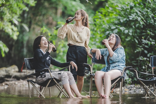 젊은 아시아 여성 그룹은 자연 공원에서 캠핑하는 동안 의자에서 맥주를 마시고 개울에 발을 담그고 함께 이야기하고 웃습니다.