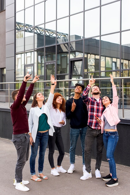 Group of young friends standing on street pointing upward in front of modern building