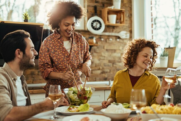 昼食を楽しんでいる若い幸せな友人のグループは、ダイニングテーブルでサラダを提供しているアフリカ系アメリカ人の女性に焦点を当てています