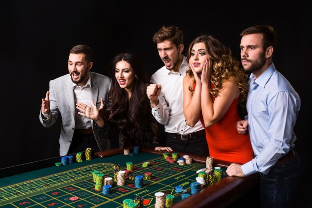 Group of young people behind roulette table on black background. Young people made bets in the game and wait for the result. Bright emotions