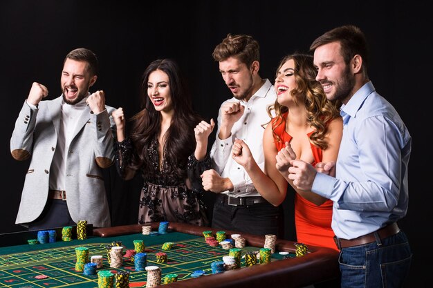 黒の背景にルーレットテーブルの後ろの若者のグループ。若い人たちはゲームに賭けて結果を待ちます。明るい感情
